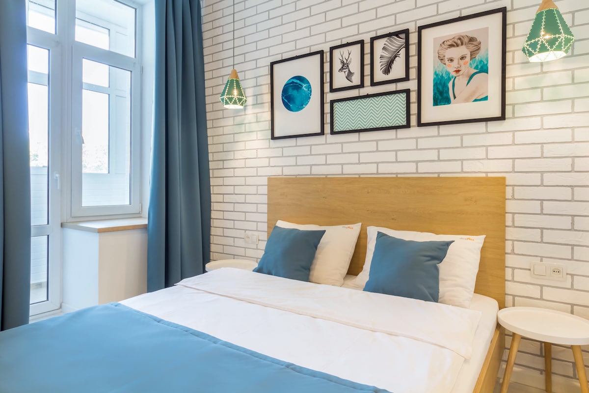 The serene sleeping area features a wooden headboard and a spacious bed with white linens and blue accent pillows. Framed artwork adorns the wall above the bed, and green pendant lights hang symmetrically. A large window with white frames allows natural light to fill the room.