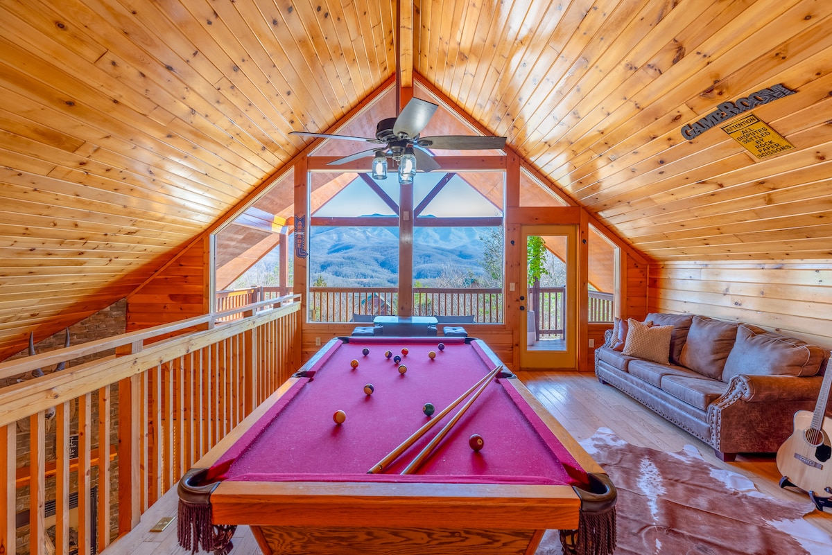 A spacious game room featuring a vibrant pink pool table positioned beneath a vaulted wooden ceiling. Large windows provide views of the surrounding mountains, while natural light fills the area. A comfortable sofa is situated nearby for relaxation.