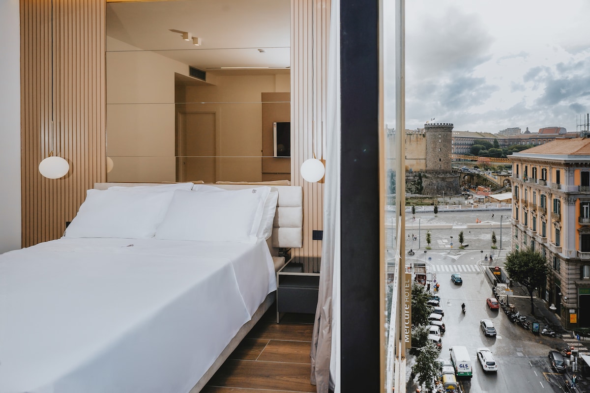 A bright sleeping area features a spacious bed with crisp white linens, set against a backdrop of large windows. Natural light filters through, offering views of a bustling street below and historic architecture in the distance.