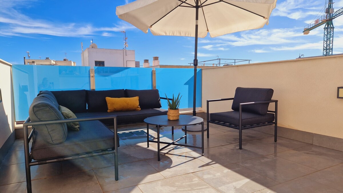 A spacious private rooftop terrace is displayed, featuring a comfortable lounge set with dark cushions and a coffee table. An umbrella provides shade, while a potted plant adds a touch of greenery. The surrounding railing is transparent, allowing for unobstructed views and ample natural light.