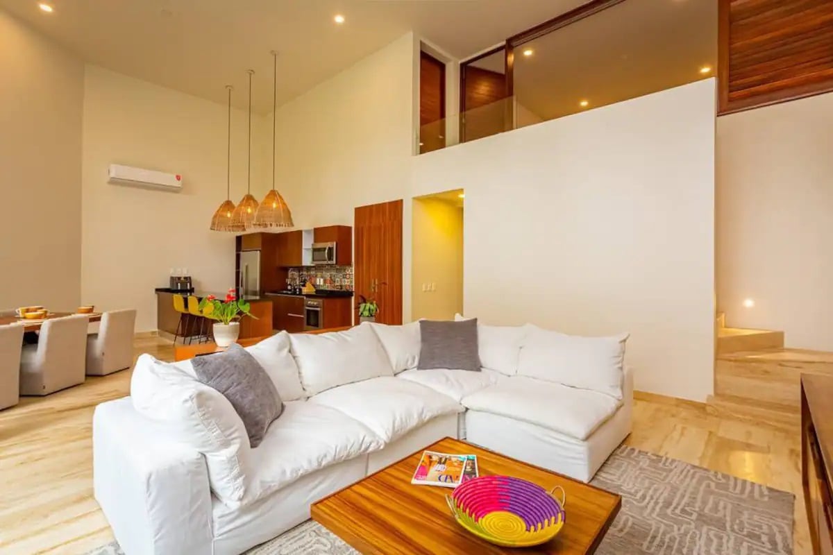 An airy living area features a large, plush sectional sofa with neutral upholstery, paired with a wooden coffee table. Pendant lighting hangs overhead, illuminating the space. The open-concept layout allows a view of the dining area, complete with modern chairs, and a kitchen in the background.