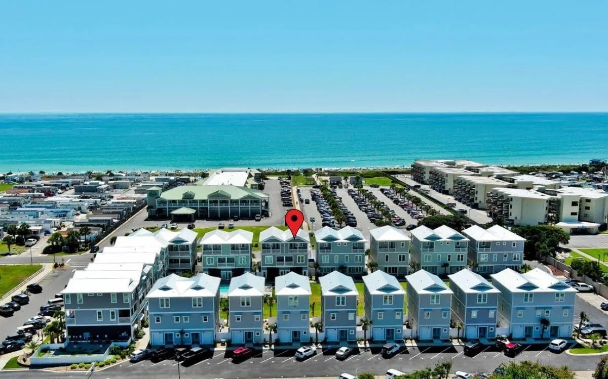 An aerial view captures the beachfront condo community with multiple modern structures lined neatly. The blue ocean and sandy beach are visible in the background, while the parking area is filled with cars. The property demonstrates proximity to both the beach and surrounding amenities.