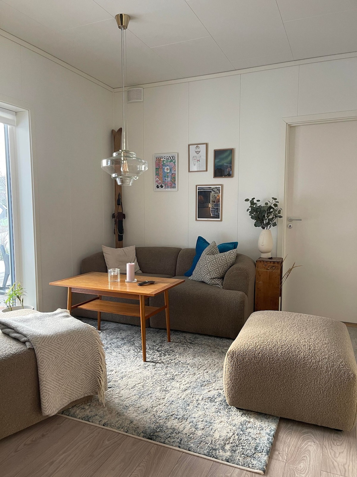 A comfortable living area is featured, with a plush sofa and a matching ottoman in soft hues. A wooden coffee table sits in the center, complemented by decorative plants and framed wall art. Natural light fills the space through a large window.