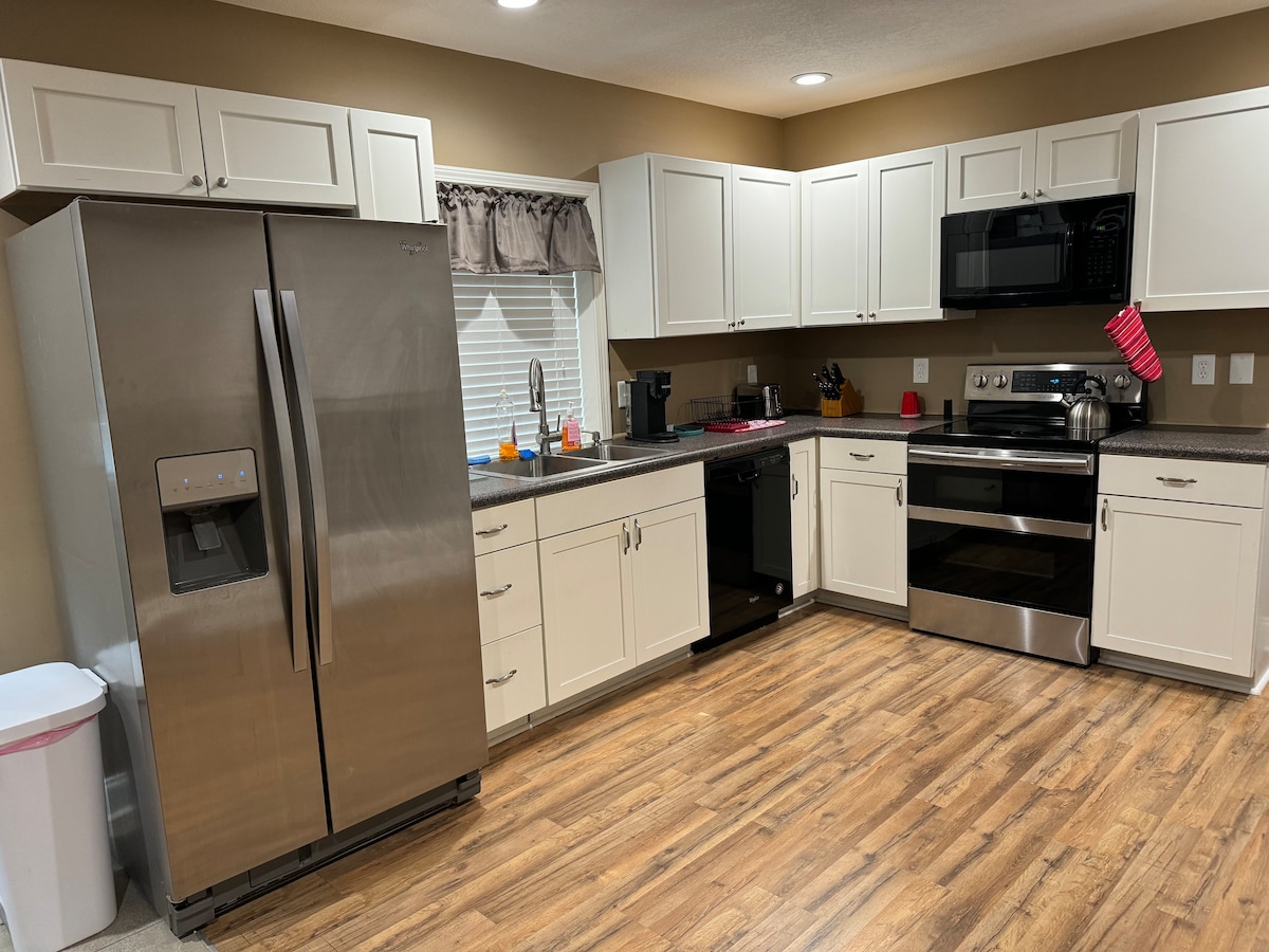 The kitchen is equipped with modern appliances, including a stainless steel refrigerator, microwave, and oven. Light-colored cabinetry complements the dark countertops, and a window offers natural light. A trash bin is discreetly placed, and wood-like flooring adds warmth to the space.
