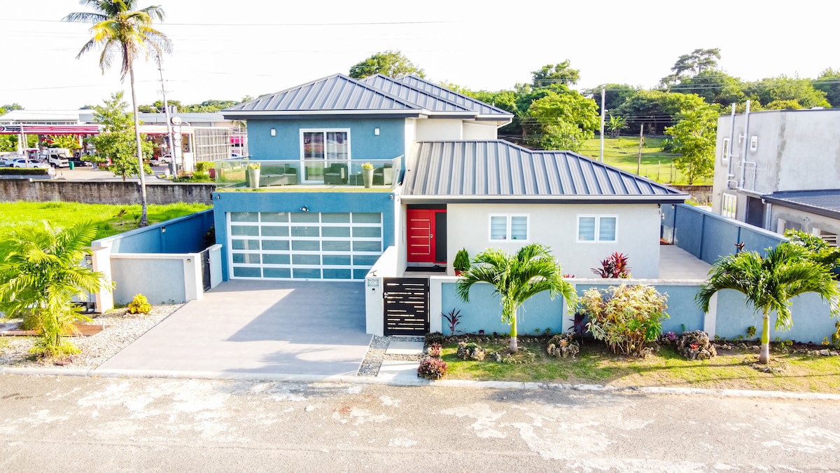 A modern two-story house is shown with a distinctive red front door. The property is surrounded by well-maintained landscaping, featuring palm trees and colorful shrubs. A spacious driveway leads to a garage with glass doors, and large windows enhance the home's contemporary charm.