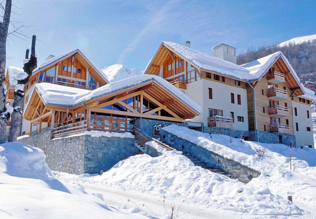 A group of chalet-style buildings is set against a snowy landscape. The design features large wooden beams and expansive windows, allowing natural light to fill the interiors. Snow covers the ground and roofs, creating a winter wonderland scene.