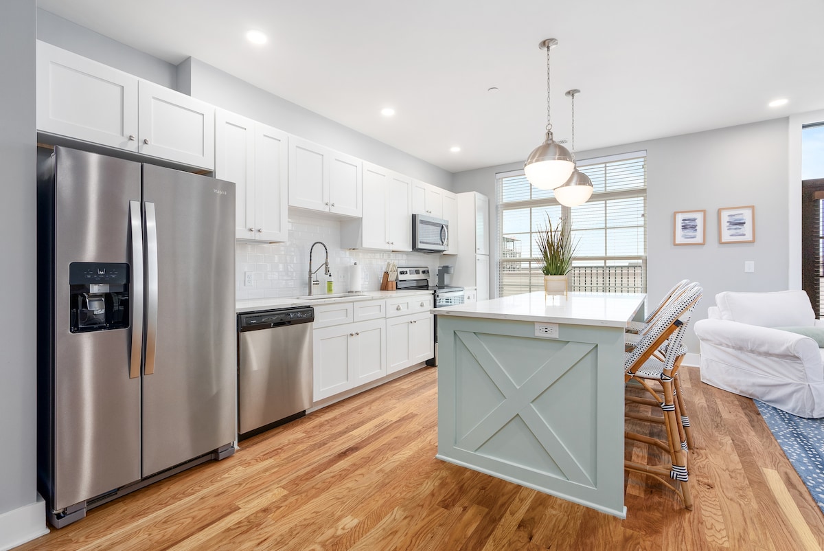 A modern kitchen features stainless steel appliances, including a refrigerator and dishwasher. White cabinetry provides ample storage, while a large island with seating invites casual dining. Natural light enters through large windows, illuminating the warm wooden flooring.