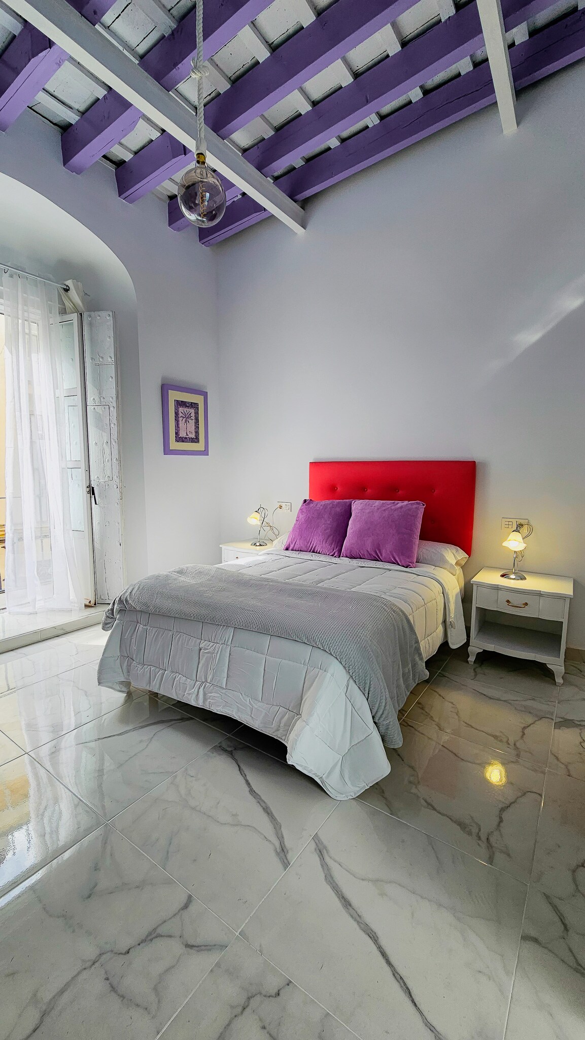 A cozy sleeping area features a vibrant red headboard with matching purple pillows on a spacious bed. Light-colored bedding complements the marble floor, while natural light filters in through a window dressed in sheer curtains, highlighting the purple-beamed ceiling.