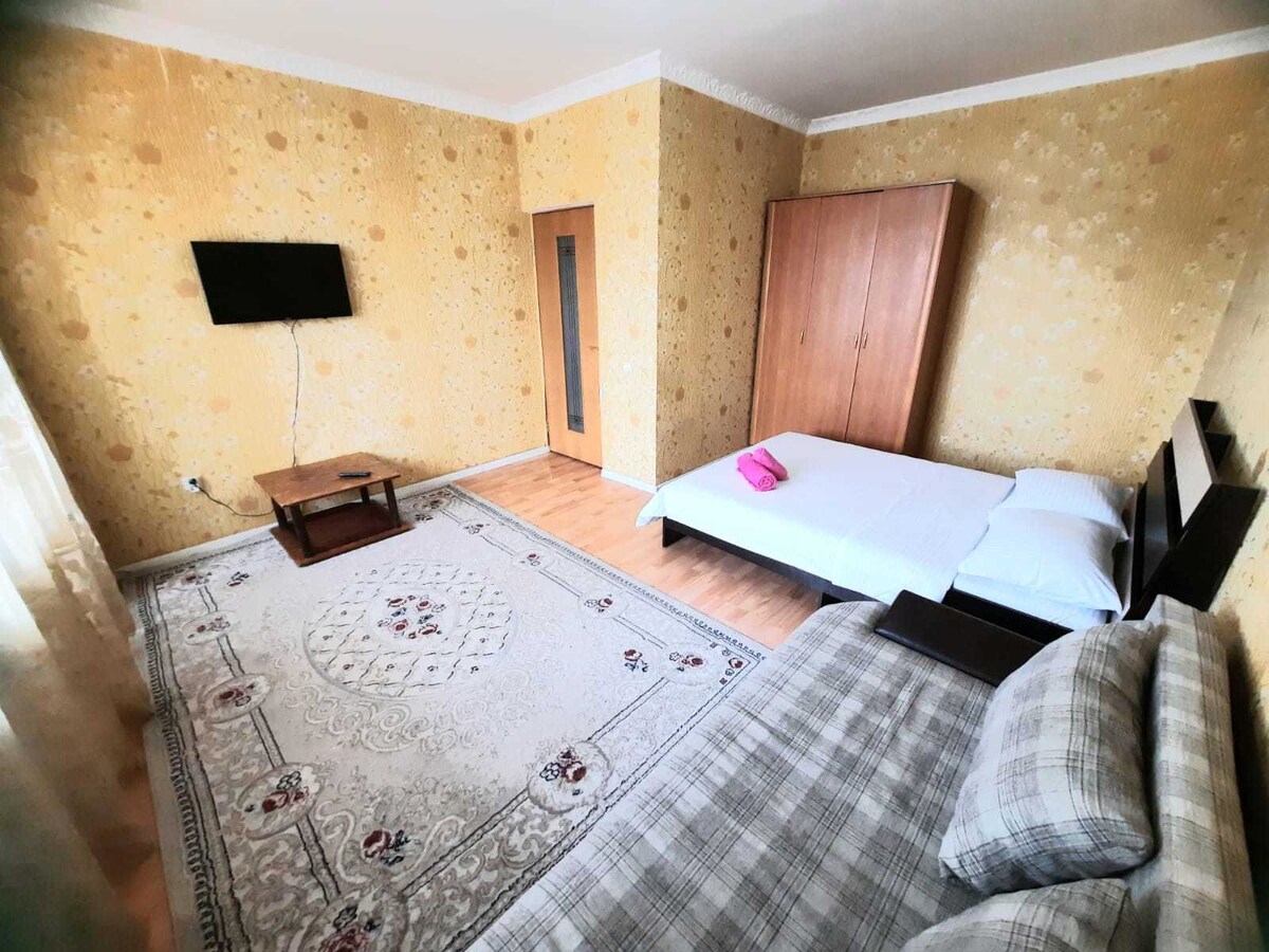 A bright bedroom features a twin bed and a double bed, complemented by a wooden wardrobe and a small coffee table. A television is mounted on the wall across from the beds. Decorative wallpaper in a light hue adds warmth to the space.