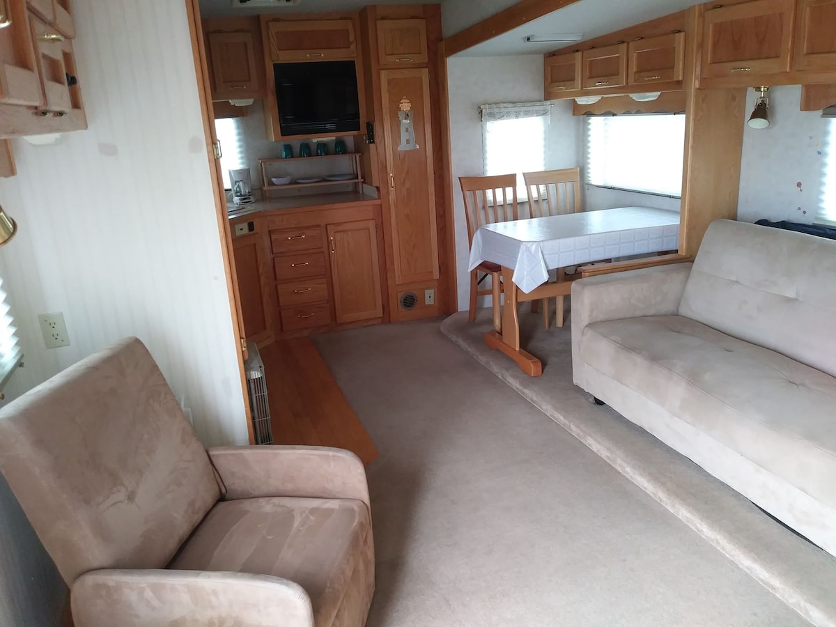 An inviting interior of the fifth-wheel reveals a cozy living area with light-colored furnishings. A table and chairs are positioned beside a window, while a compact kitchen area features wooden cabinetry. Soft carpeting enhances the warmth of the space, allowing for comfortable relaxation.