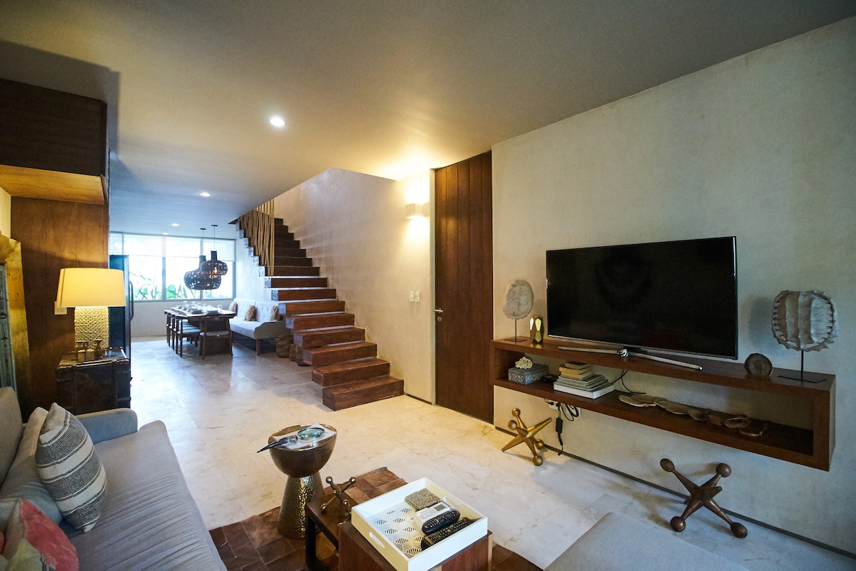 A spacious living area showcases modern furnishings and a neutral color palette. Soft lighting creates a warm ambiance, and a large television is positioned on a wooden shelf. A staircase leads to the upper levels, enhancing the open layout of the home.