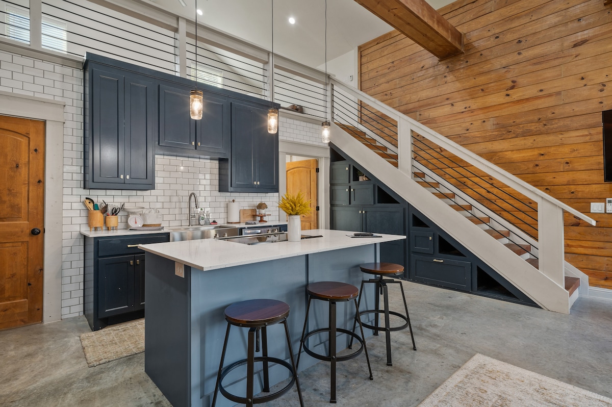 A modern kitchen is showcased with navy blue cabinetry and white subway tiles. An island with seating for three is present, complemented by pendant lighting. Exposed wooden beams and a staircase enhance the open concept design, while a warm color palette adds to the inviting ambiance.