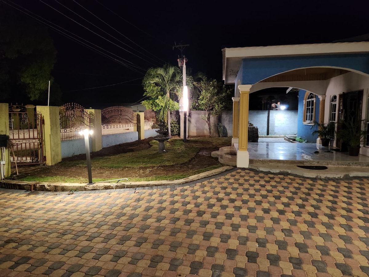 An outdoor space is illuminated by soft lighting at night, showcasing a paved driveway with a checkerboard pattern. The area is bordered by well-kept greenery and a fence. A building with vibrant blue and yellow accents is visible to the right.