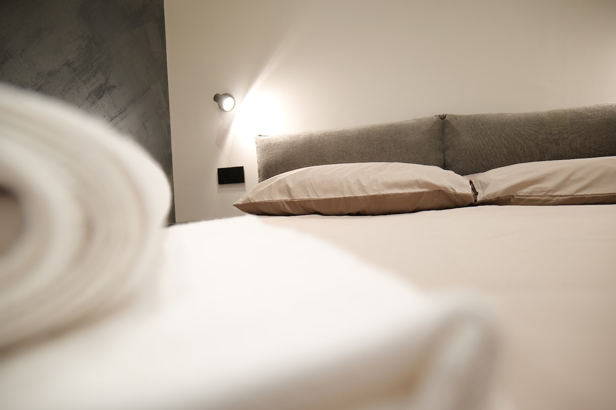 A spacious bed with soft, neutral bedding is featured, positioned against a minimalist wall. A modern light fixture illuminates the area, while neatly folded linens are prominently displayed in the foreground.