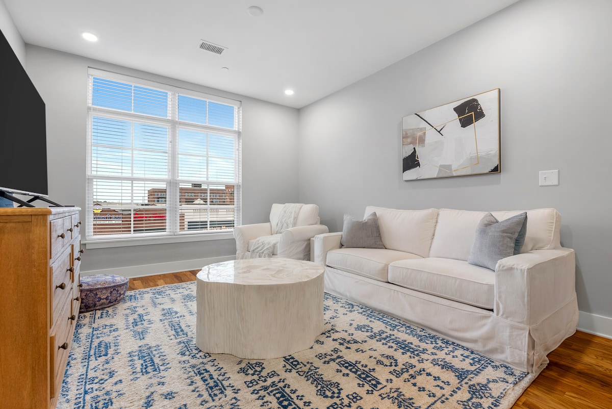 A bright and spacious living area features a large white couch and a comfortable armchair. A round wooden coffee table sits on a decorative area rug. Light streams in through large windows, enhancing the open and airy feel of the room.