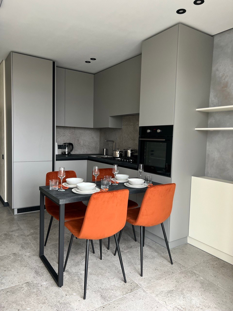 A modern kitchen area features sleek cabinetry and a dark countertop. A small dining table with four orange chairs is set with dishes and glassware. An oven and coffee maker are visible within the cabinetry, creating a functional and stylish space.