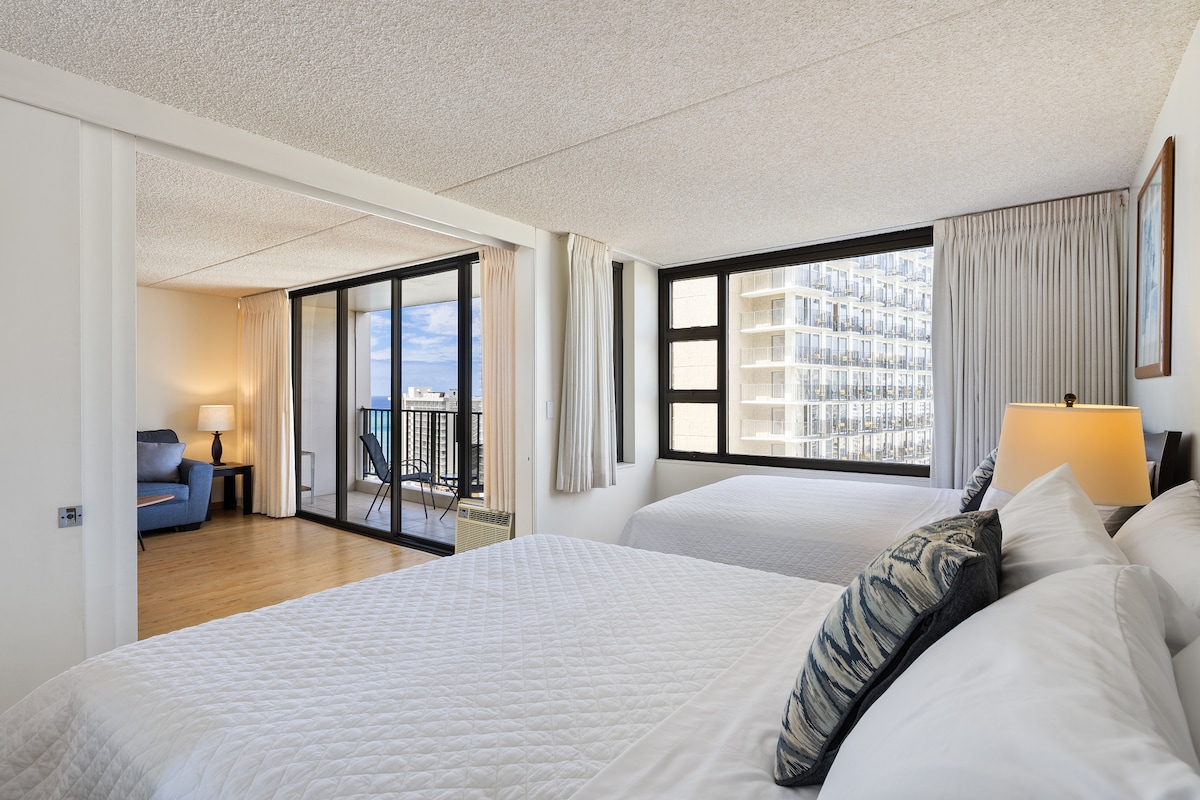 The image showcases a spacious bedroom featuring two queen beds with crisp white bedding. Large windows allow natural light, revealing a view beyond. A comfortable seating area is visible, with a blue chair next to a lamp, and sliding glass doors provide access to a private balcony.