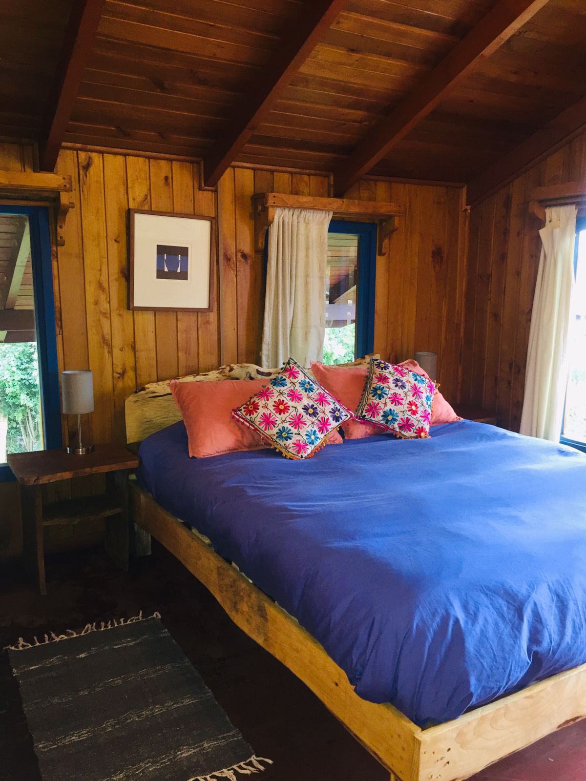 A cozy sleeping area features a spacious bed with blue bedding and colorful decorative pillows. Natural light filters through the windows, illuminating the wooden walls and ceiling. A small lamp and a rug add warmth to the welcoming atmosphere.
