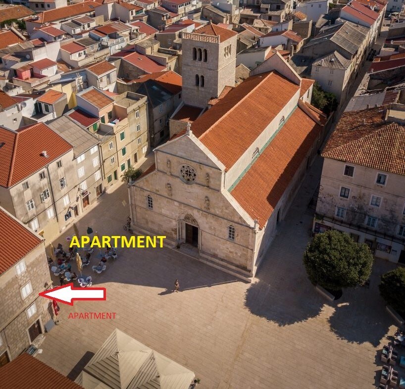 An aerial view showcases the apartment's location adjacent to a historic church amidst the main square of Pag. Surrounding buildings feature a variety of roof colors, and outdoor seating is visible on the plaza, conveying a vibrant atmosphere.