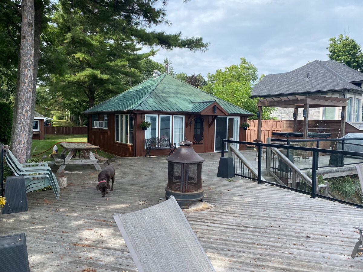 The cabin features a unique circular shape with a green metal roof, surrounded by trees. A spacious wooden deck extends toward the waterfront, showcasing seating areas, a fire pit, and a nearby dog exploring the space. A glimpse of the water and neighboring houses can be seen.