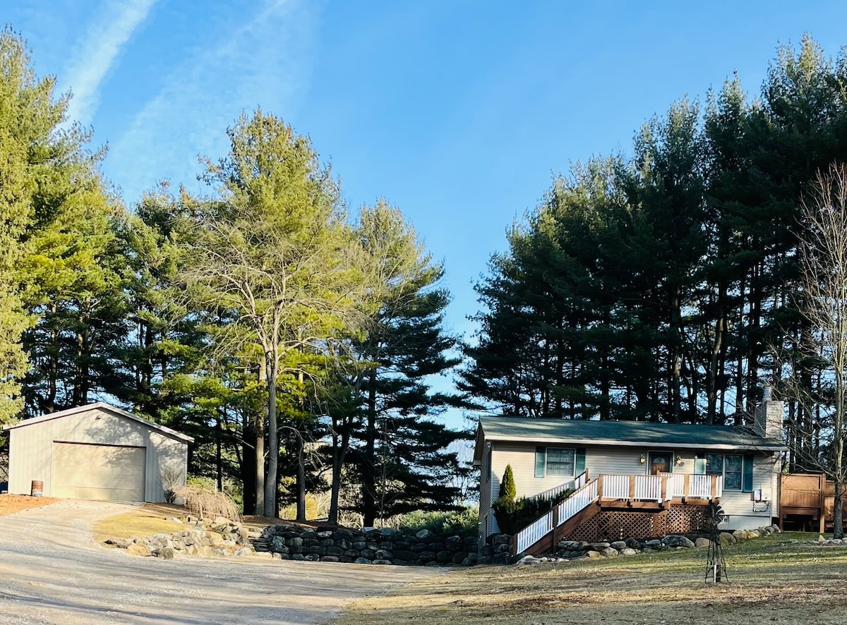 A ranch-style home is shown set in a serene countryside landscape, surrounded by tall evergreen trees. A gravel driveway leads to the house, which features a wraparound deck. A spacious outbuilding can be seen nearby, completing the tranquil setting.