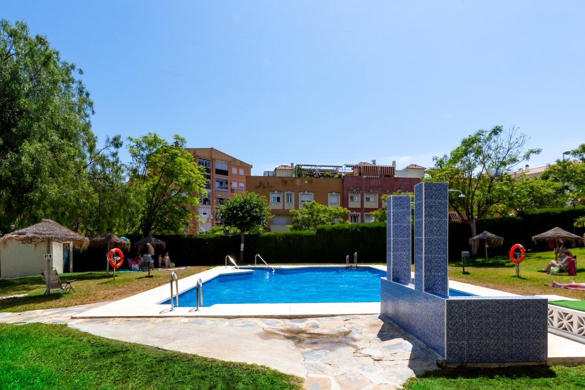 A communal swimming pool is surrounded by a grassy area with sun loungers and shaded cabanas. The pool features clear blue water and is complemented by nearby loungers, creating a space for relaxation. Lush trees provide a natural backdrop to the scene.