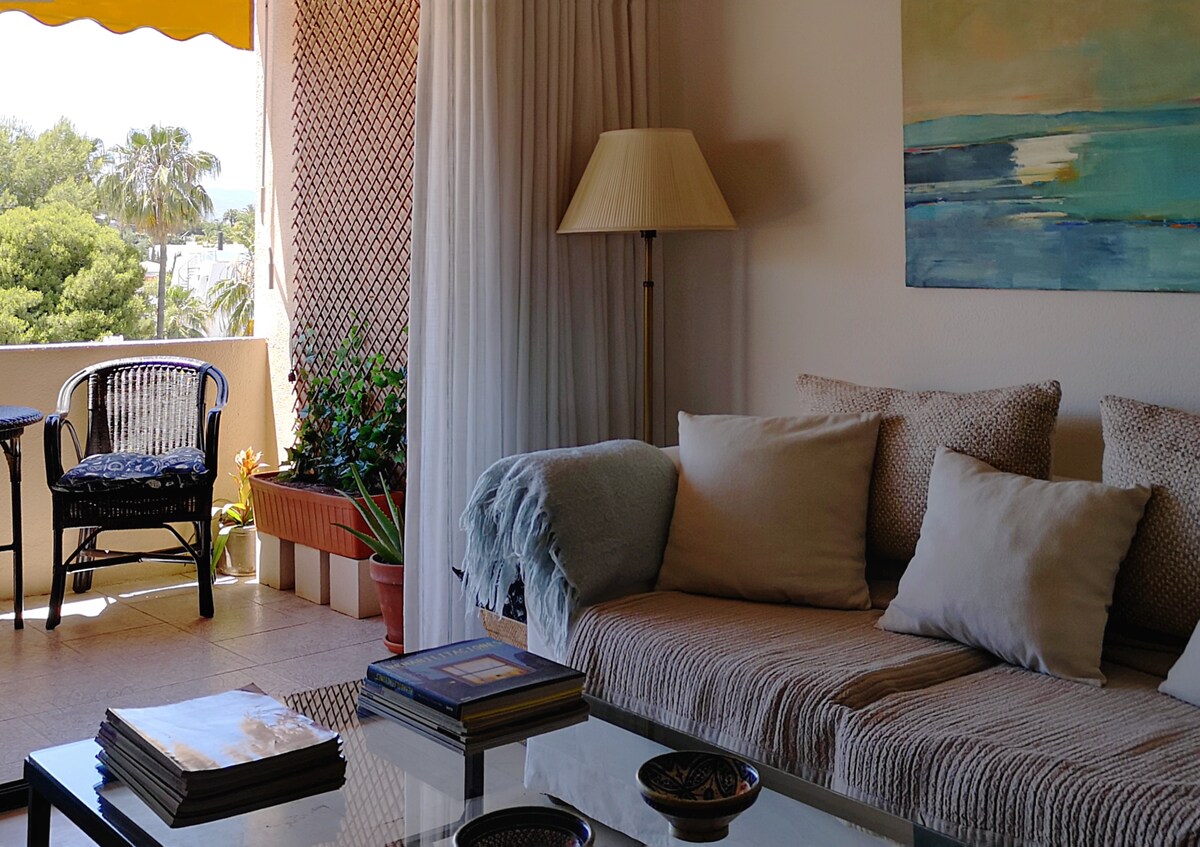 A comfortable sitting area is presented with a cozy sofa adorned with light pillows. A glass coffee table is positioned in front of the sofa. Nearby, a balcony is visible, featuring a pair of cane chairs under a yellow awning, with greenery in the background.