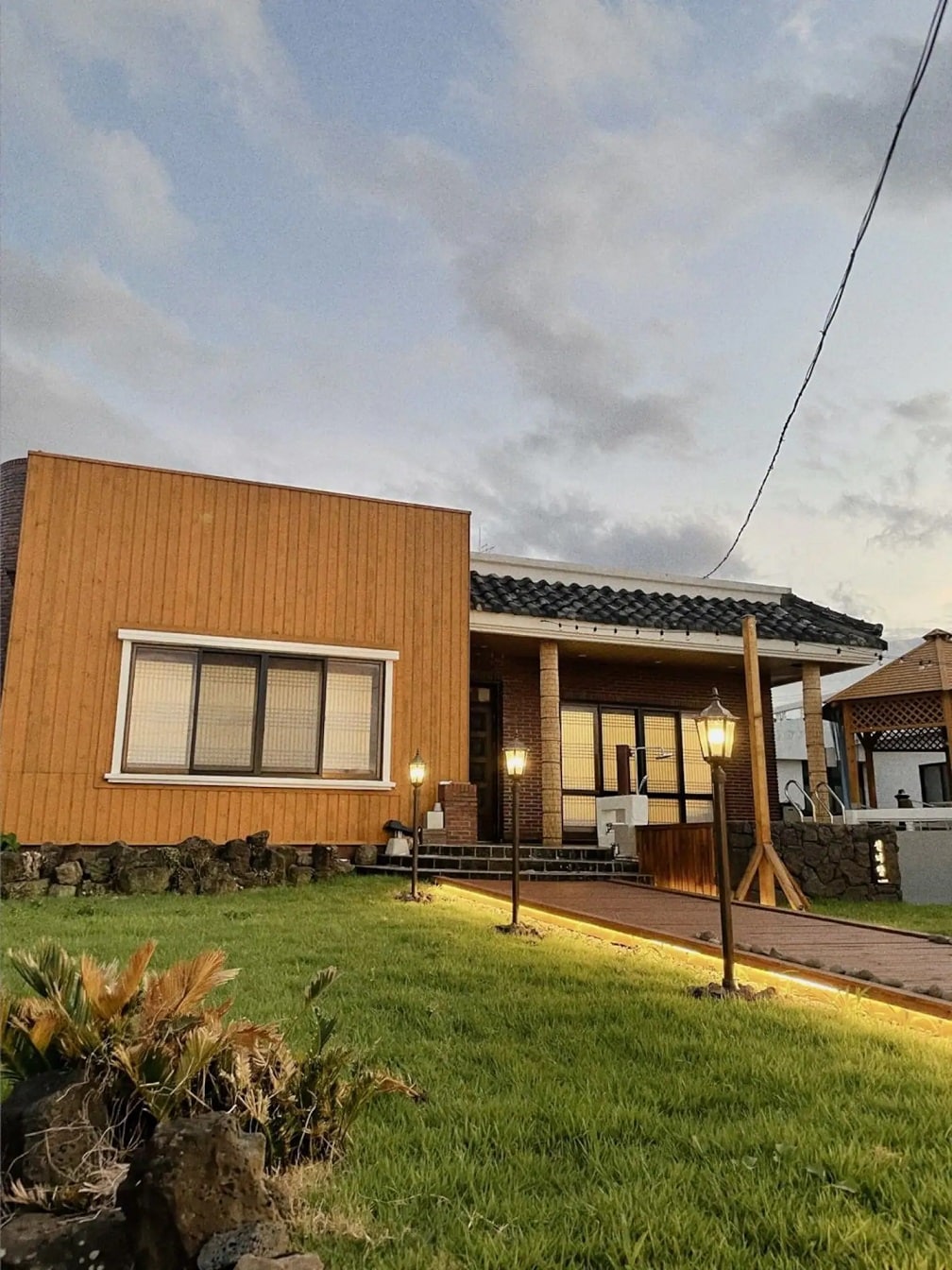 The exterior of the accommodation features a warm wooden faรงade complemented by a stone pathway illuminated by decorative lamps. A spacious porch offers access to the entrance, while lush green grass surrounds the area, creating a serene environment.