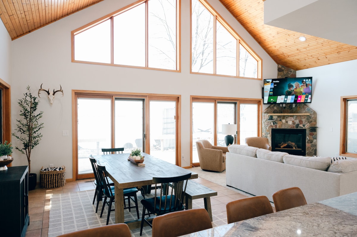 A bright and airy living space is showcased with large windows allowing natural light to flood the room. A spacious dining table is set beside a cozy seating area, where a stone fireplace adds a welcoming touch. A large television is mounted on the wall, enhancing the modern ambiance.