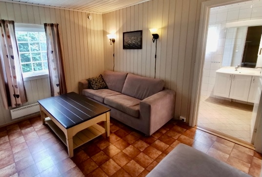 A light-filled seating area features a comfortable couch and a coffee table, set against wooden paneling. Natural light enters through a window adorned with soft curtains. A doorway leads to a tiled bathroom visible in the background.