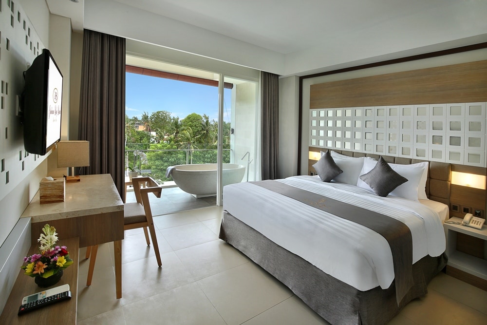 A spacious room is presented, featuring a large bed adorned with crisp white linens. A wooden desk and chair are situated beside a flat-screen TV. Floor-to-ceiling glass doors open to a balcony with a bathtub, overlooking lush greenery.