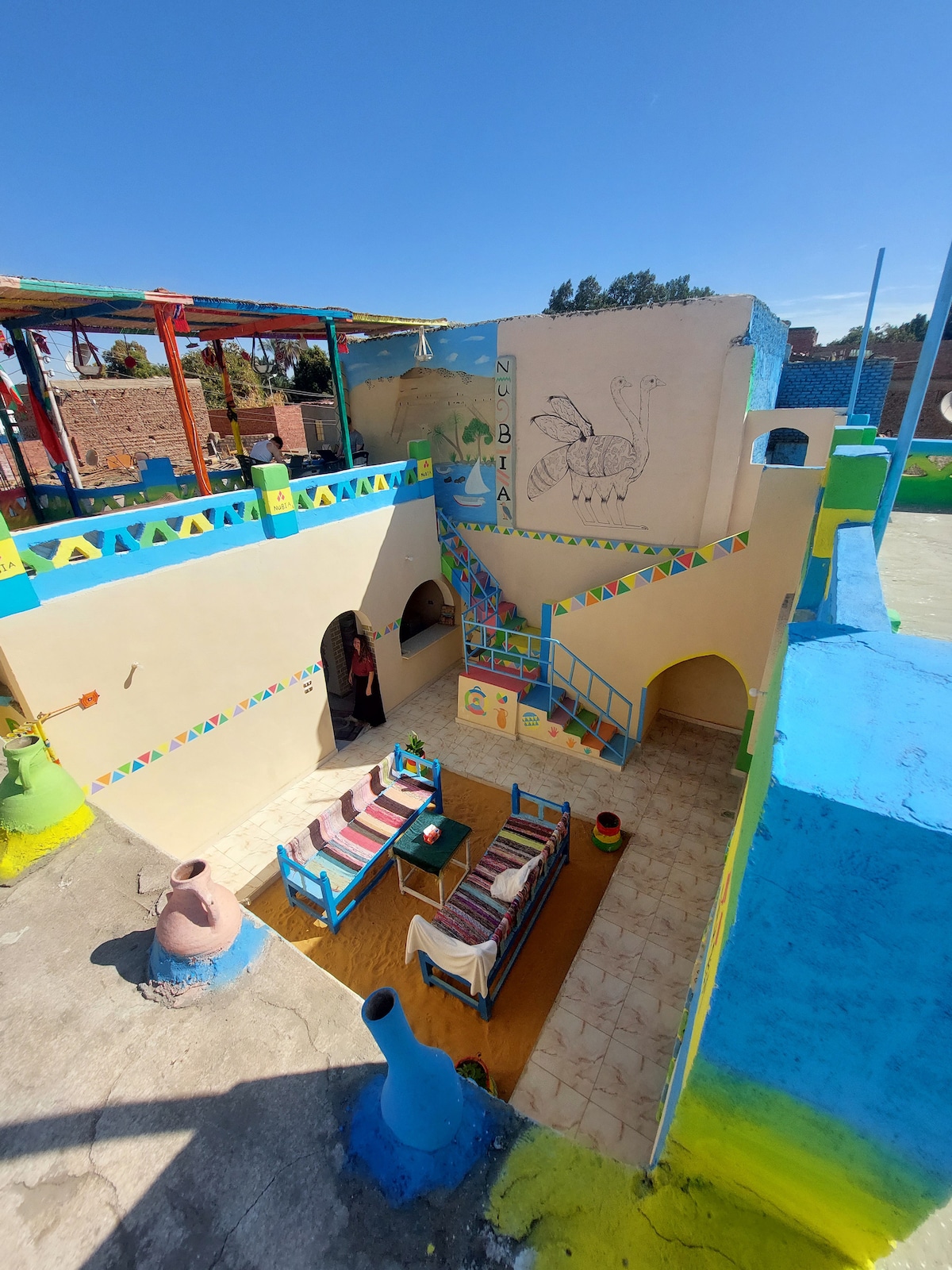 A vibrant courtyard is showcased, featuring colorful walls in shades of blue and yellow. A seating area with striped cushions is positioned on the tiled floor, accompanied by decorative pottery. A staircase leads to an upper level, enhancing the layered appearance of the space.