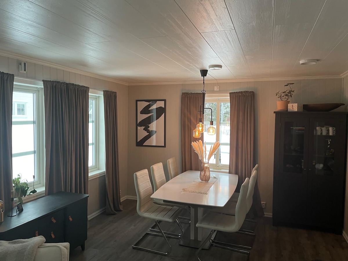 A dining area features a large, white table surrounded by six sleek white chairs. Natural light enters through two windows, with brown curtains providing a soft backdrop. A dark cabinet stands against one wall, complemented by a framed artwork that adds a touch of style.