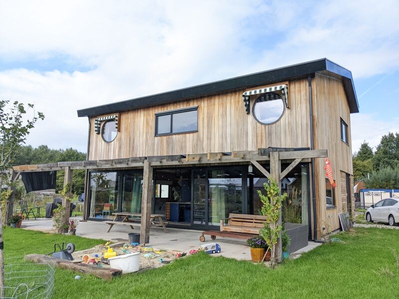 The modern two-story home is constructed from natural materials, featuring large glass panels and round windows. An expansive outdoor space includes a concrete patio with seating and play areas. Lush green grass surrounds the house, and plants are positioned around the entry, enhancing the environment.