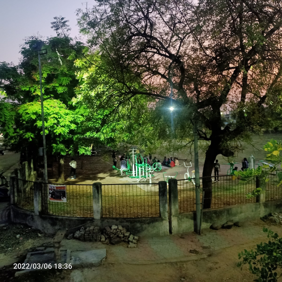 An outdoor fitness area is shown, surrounded by a fence and illuminated by nearby lights. Green exercise equipment is visible, along with trees that provide shade. Several individuals can be seen engaging in physical activities, creating a vibrant community atmosphere.