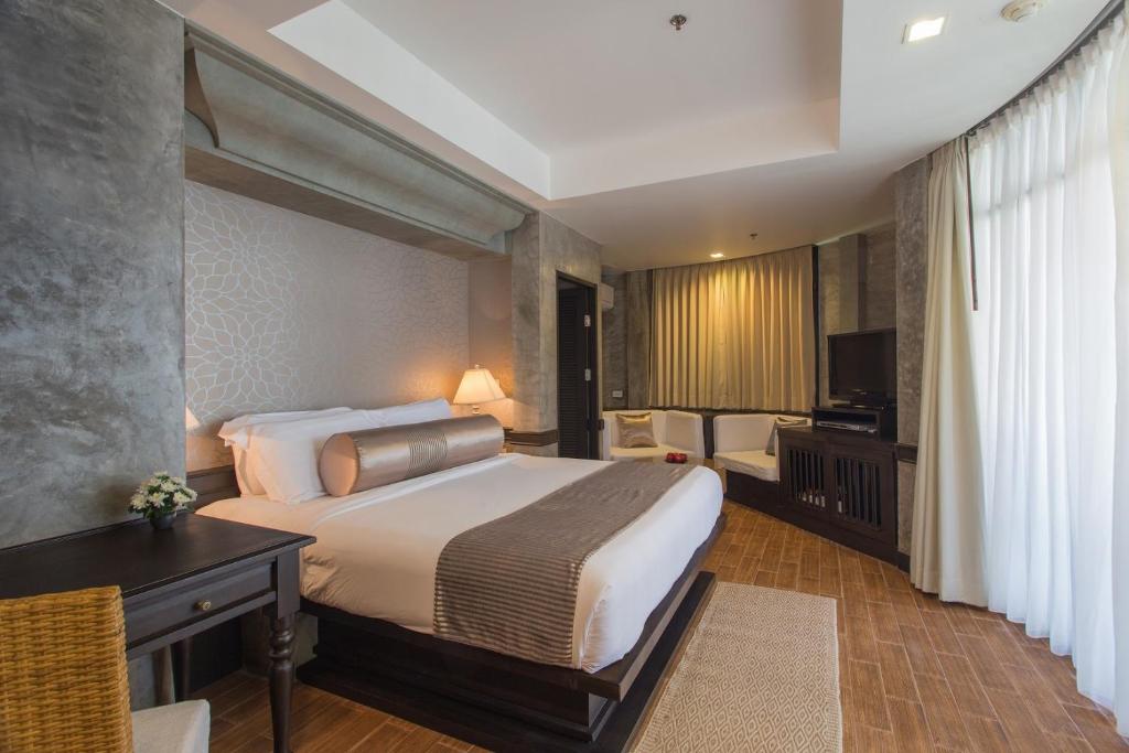 A well-furnished room features a spacious bed with soft linens, complemented by muted tones. A comfortable seating area includes a chair and small table. Natural light flows in through floor-to-ceiling curtains, and a television is positioned on a dark wooden unit.