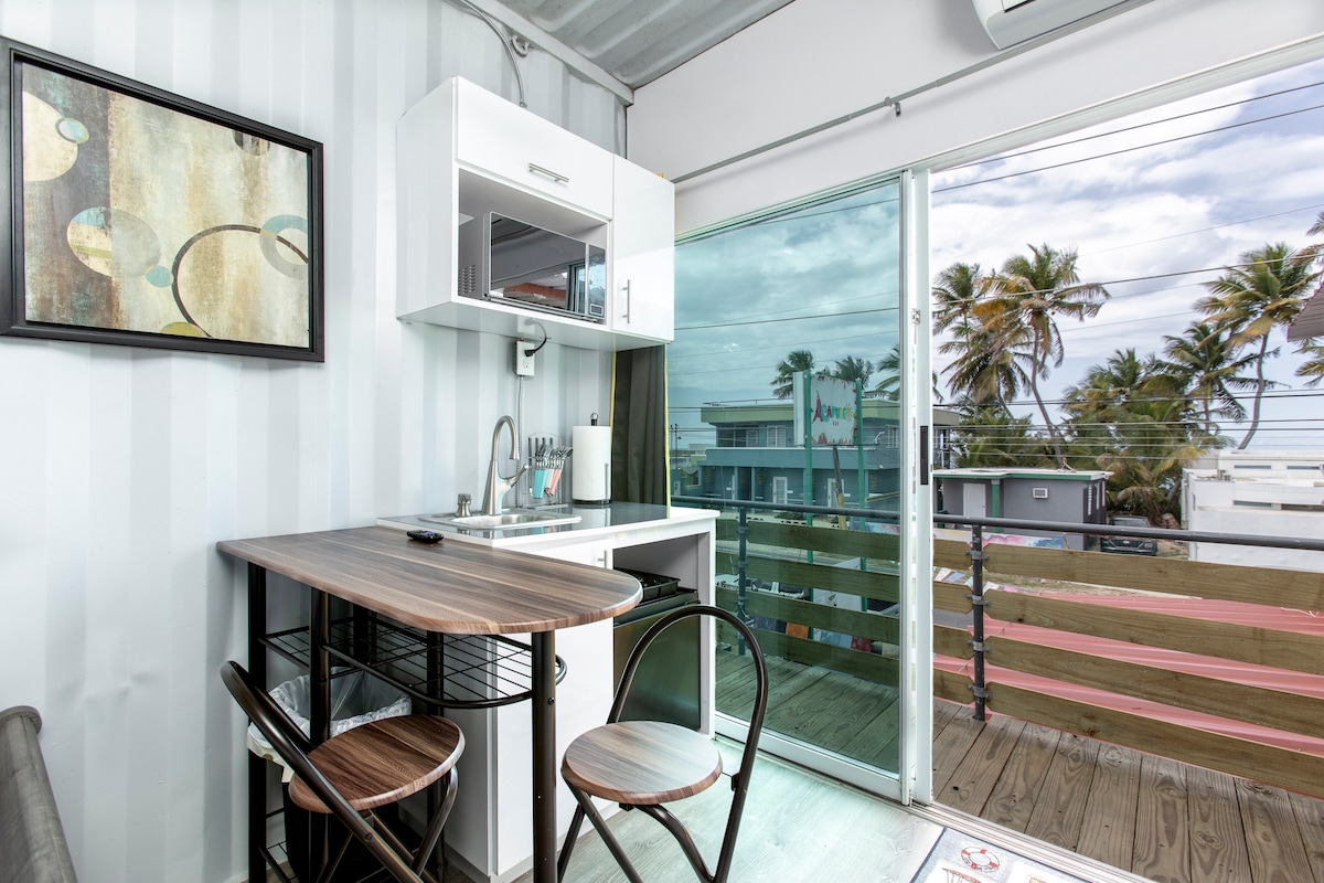 A compact dining area features a small table and two chairs against a backdrop of large glass doors. Natural light filters in, illuminating a minimalist kitchen setup with cabinetry and a sink. Tropical palm trees are visible outside the glass doors.