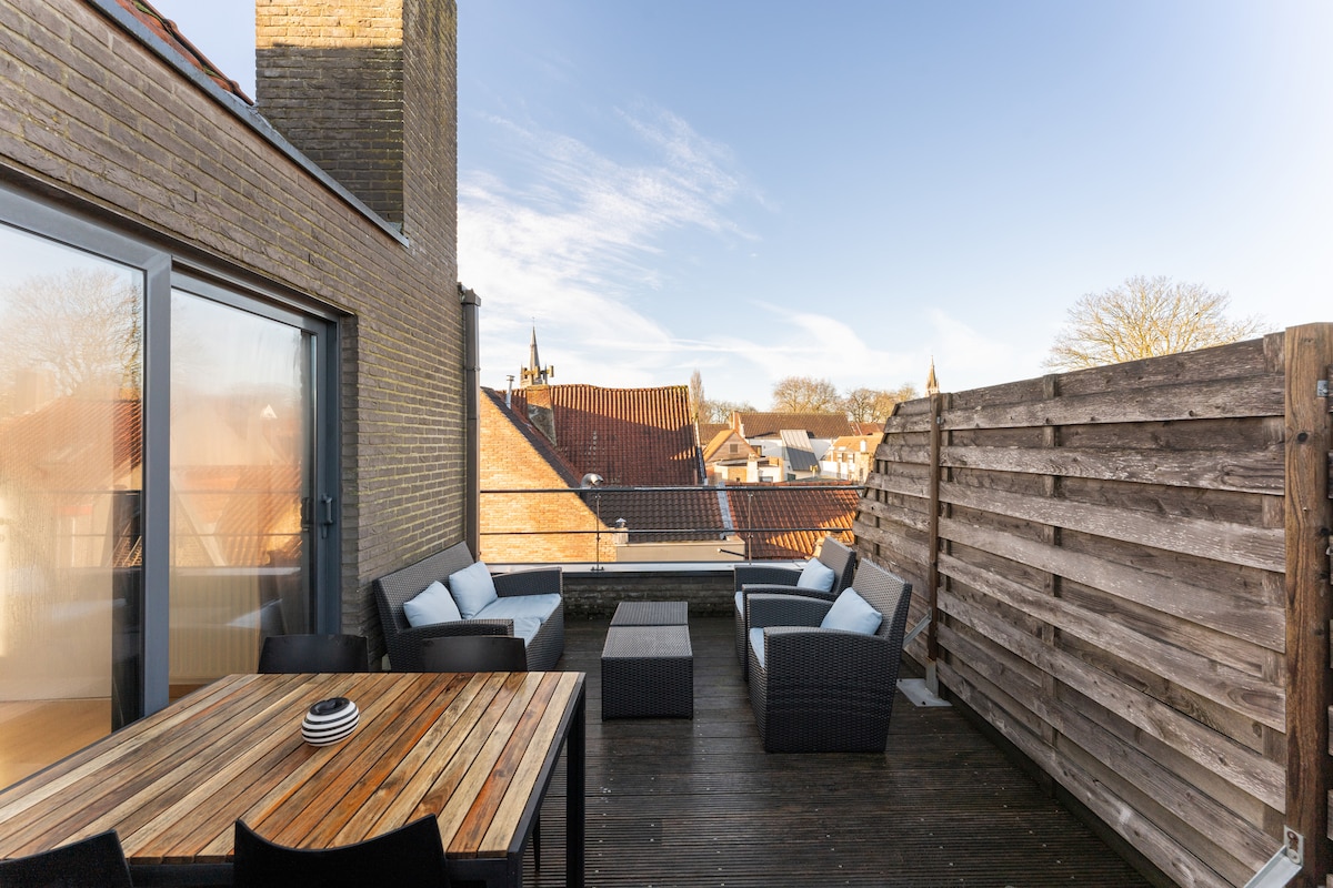 A spacious sun terrace is presented with comfortable seating arrangements and a dining table. Surrounding views of rooftops and iconic towers of Brugge are visible. The terrace offers a private atmosphere, enhancing outdoor relaxation.