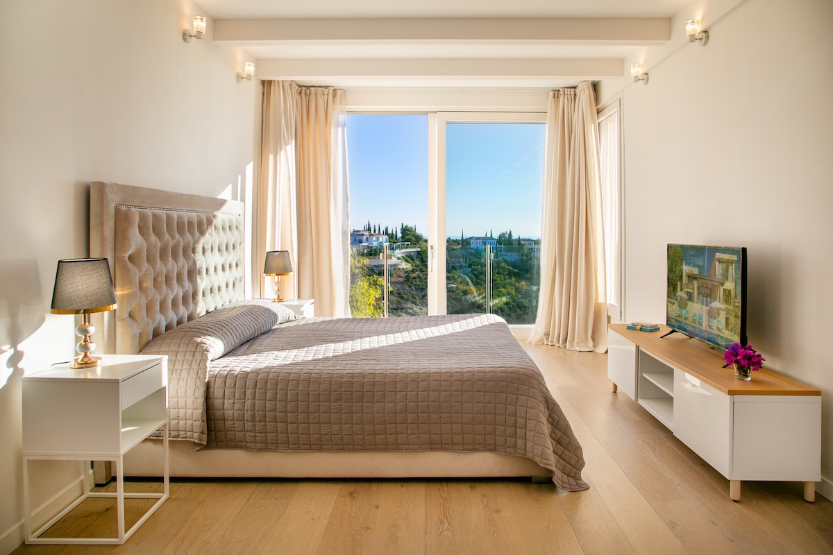 A bright and spacious bedroom features a comfortable bed with a neutral quilted cover. Modern furniture includes two bedside tables and a media console. Large windows allow natural light to fill the space, offering scenic views of the surrounding landscape.