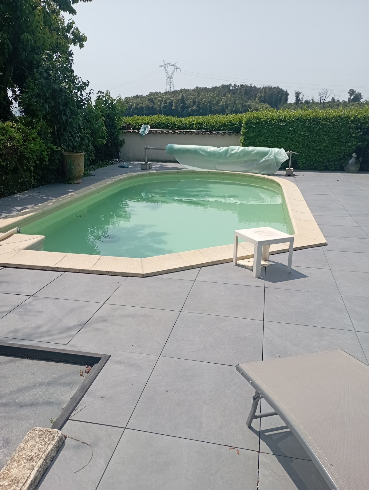 A rectangular pool is surrounded by a light-colored stone deck, featuring a small side table and a sun lounger. Lush greenery forms the backdrop, and an expansive view of the landscape is visible in the distance.
