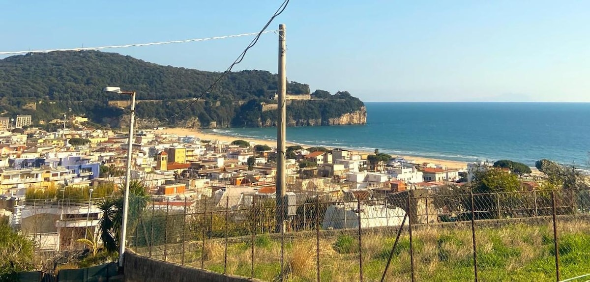 A panoramic view captures a sweeping landscape of the coastline and city below. The beach is visible, along with a cluster of colorful buildings. Rolling hills rise in the background, while the calm sea stretches into the distance under clear skies.