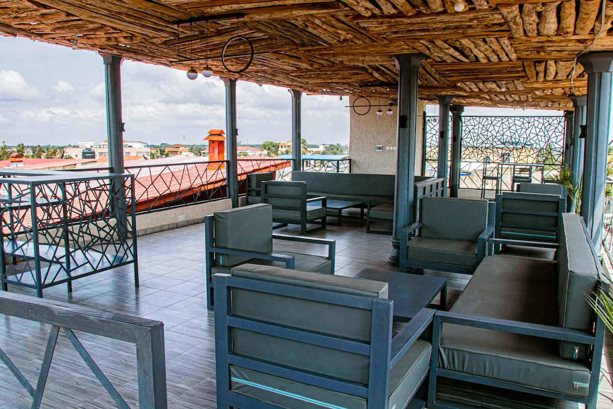 A spacious rooftop area features several seating arrangements with modern gray sofas and a dark table. The ceiling showcases natural wooden beams, while a decorative guardrail provides a view of the surrounding area. Soft lighting fixtures add to the ambiance.