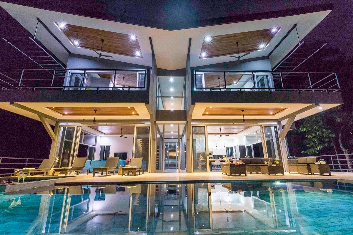 The modern two-story villa is illuminated at night, showcasing sleek architectural lines. Large windows reflect the surrounding environment, while a spacious outdoor area is furnished with lounge chairs. A serene pool mirrors the building's design, enhancing the overall visual appeal.
