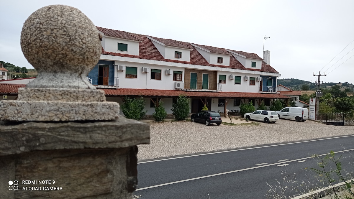 The low-rise building is set back from the road, with a gravel parking area in front. Multiple air conditioning units are visible on the façade. Nearby, a row of trees provides a natural landscape, while vehicles are parked neatly along the driveway.