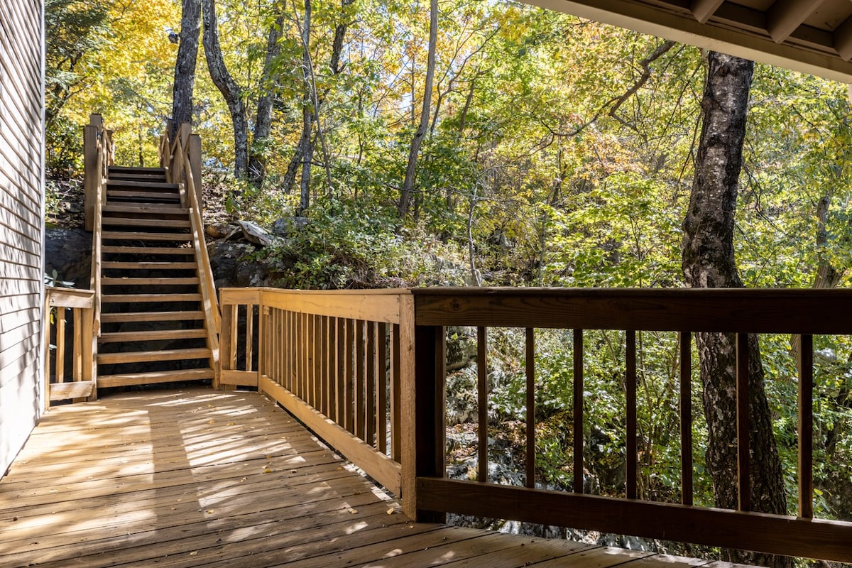 A wooden deck connects to a pathway lined with trees displaying vibrant autumn foliage. Natural light filters through the leaves, casting gentle shadows on the steps leading upwards, inviting exploration of the surrounding forest.