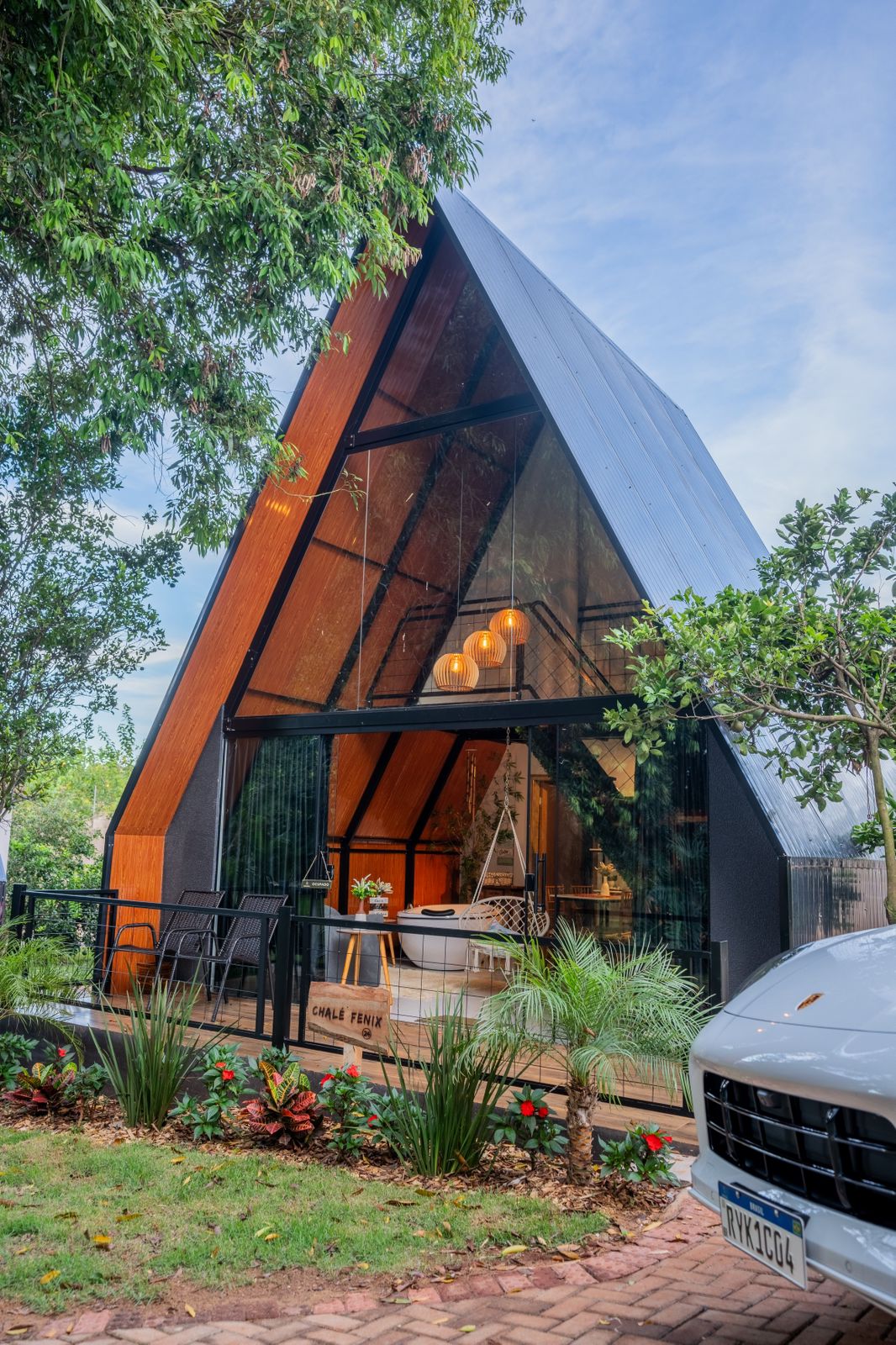 The cabin features a striking triangular architecture with large glass panels, allowing sunlight to illuminate the interior. Surrounding greenery and carefully arranged plants enhance the serene entrance, while a cozy porch with seating invites relaxation.