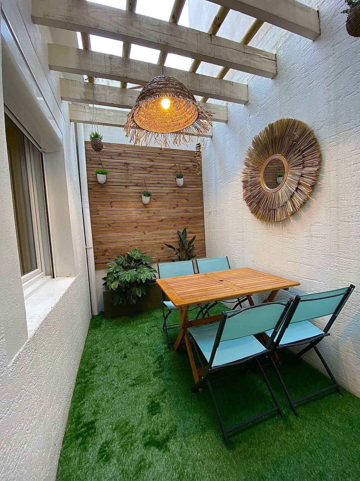 An outdoor patio space is featured, featuring a wooden table surrounded by four folding chairs with a light blue finish. Decorated with green plants and a wooden wall art piece, the area also includes a pendant light hanging from a pergola above, enhancing the tranquil setting.