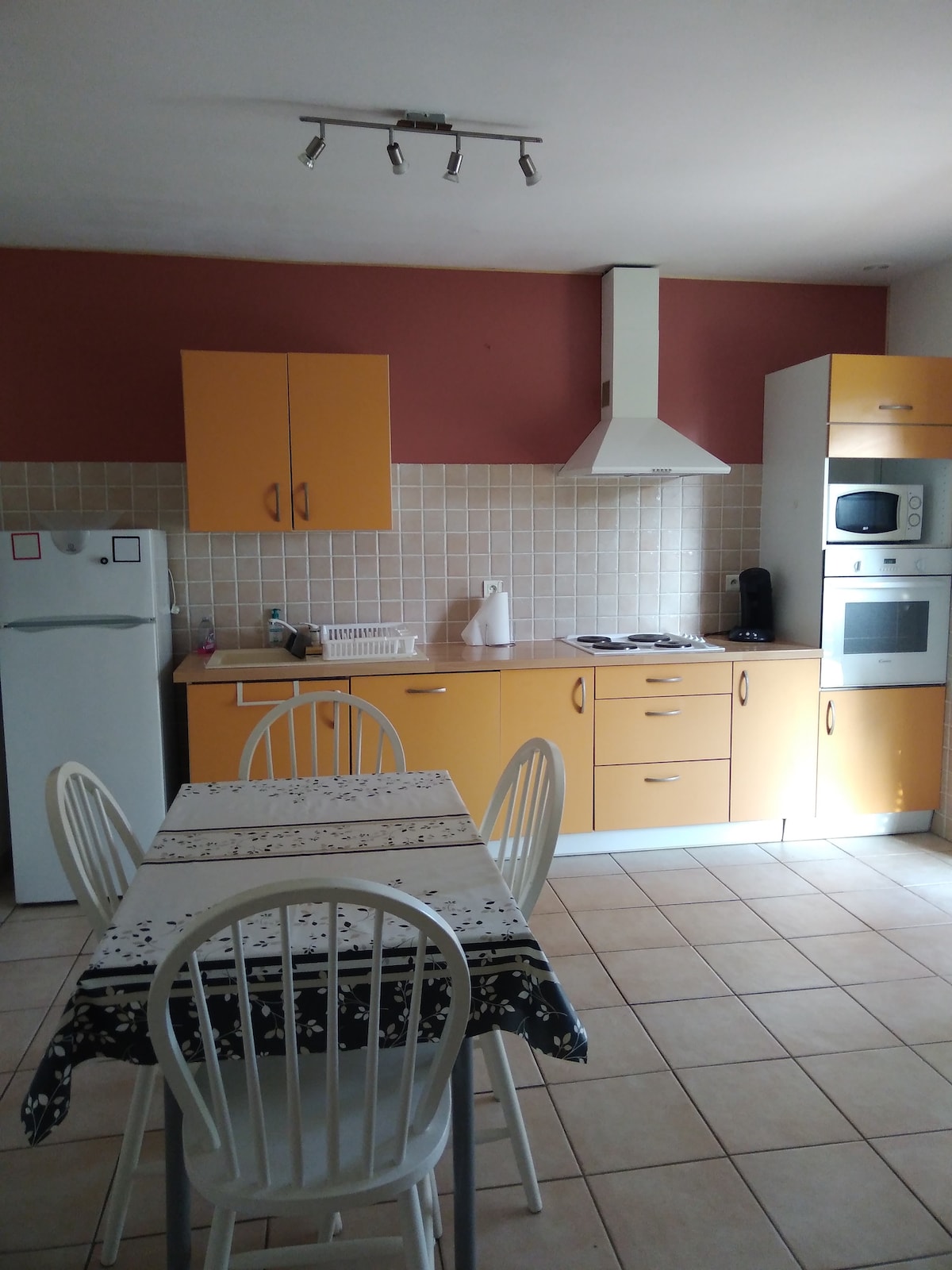 The kitchen features orange cabinetry and a light tile floor. A dining table with four white chairs is positioned in the center. Essential appliances, including a refrigerator, stove, and microwave, are situated along the wall, complemented by a neutral tiled backsplash.
