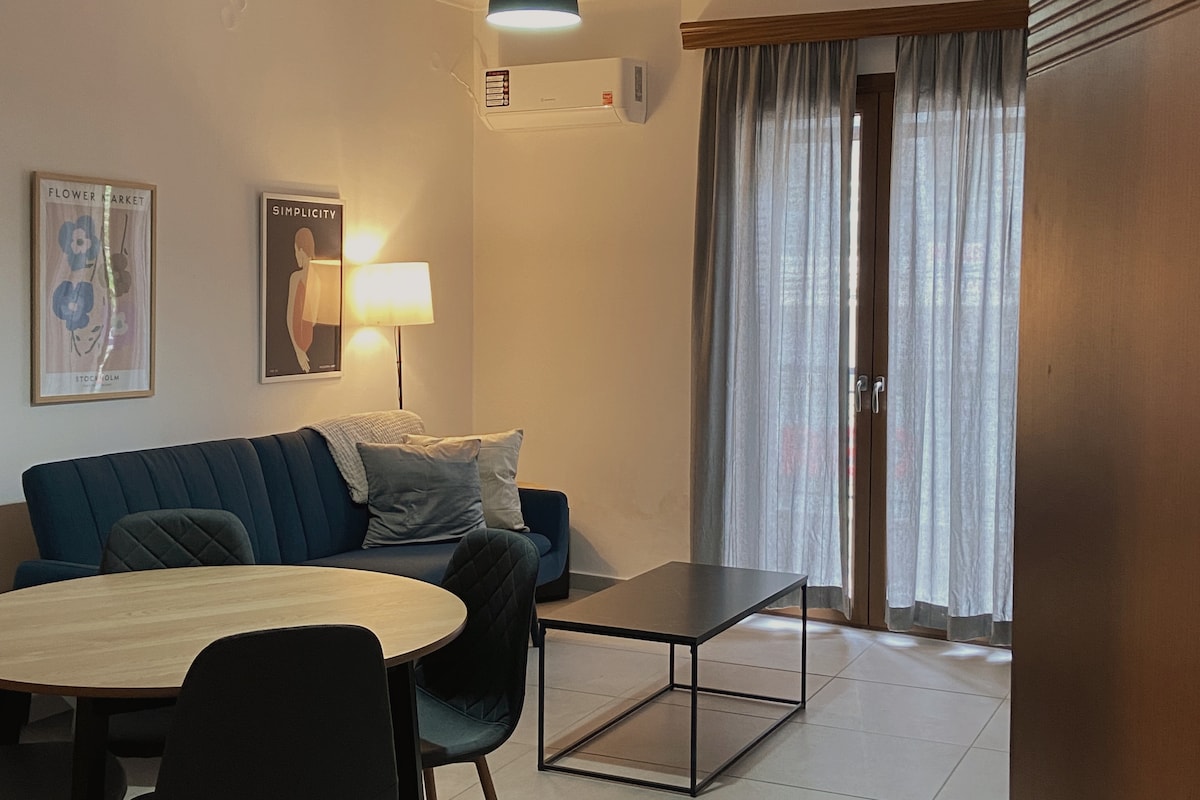A modern living space features a deep blue sofa adorned with decorative pillows. A round dining table is paired with black chairs, while a sleek coffee table occupies the center. Natural light filters through sheer curtains, and an airy ambiance is created by the subtle lighting.
