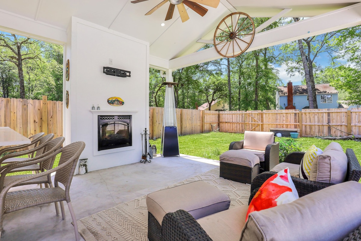 A spacious outdoor living area is equipped with comfortable seating arranged around a fire feature, under a ceiling fan. A dining table accommodates several chairs, and the space is surrounded by greenery and a wooden fence, creating a private atmosphere.